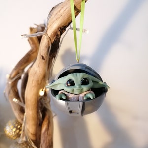 May include: A green and gray Christmas ornament featuring a character from the Star Wars franchise, The Mandalorian. The ornament is shaped like a pod and the character is sitting inside. The ornament is hanging from a green ribbon.