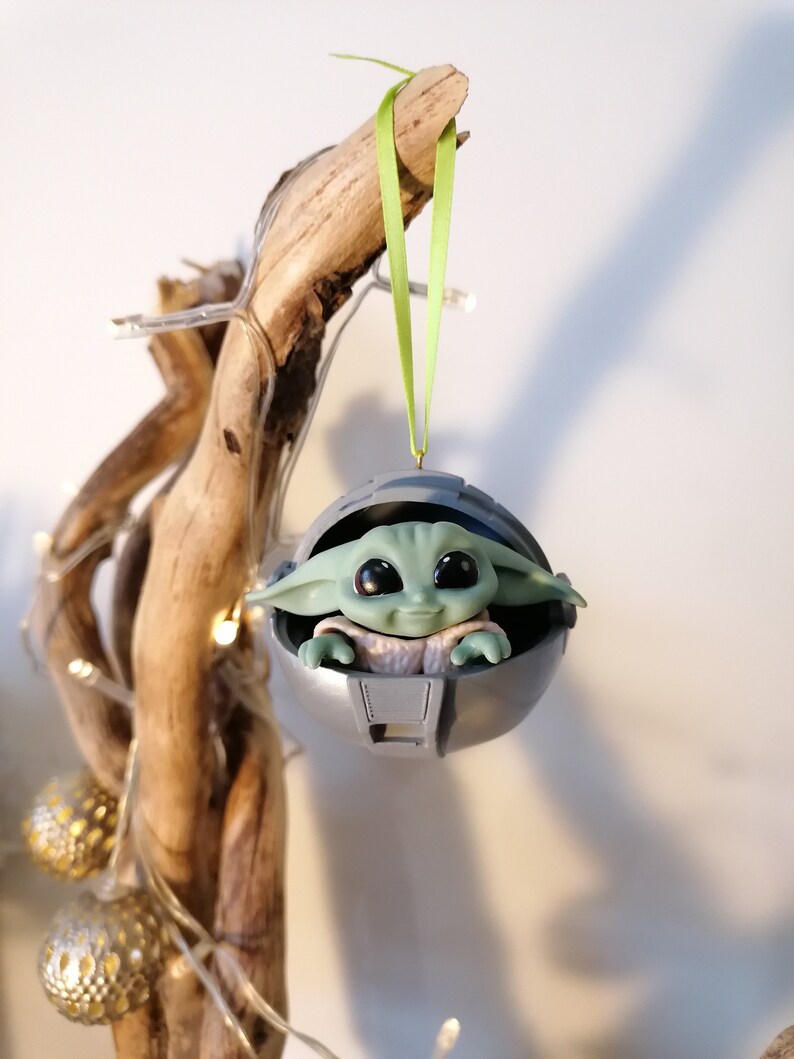 May include: A green and gray Christmas ornament featuring a character from the Star Wars franchise, The Mandalorian. The ornament is shaped like a pod and the character is sitting inside. The ornament is hanging from a green ribbon.