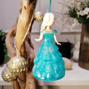 May include: A blue and white plastic ornament of a Disney princess wearing a sparkly blue dress. The ornament is hanging from a blue ribbon.