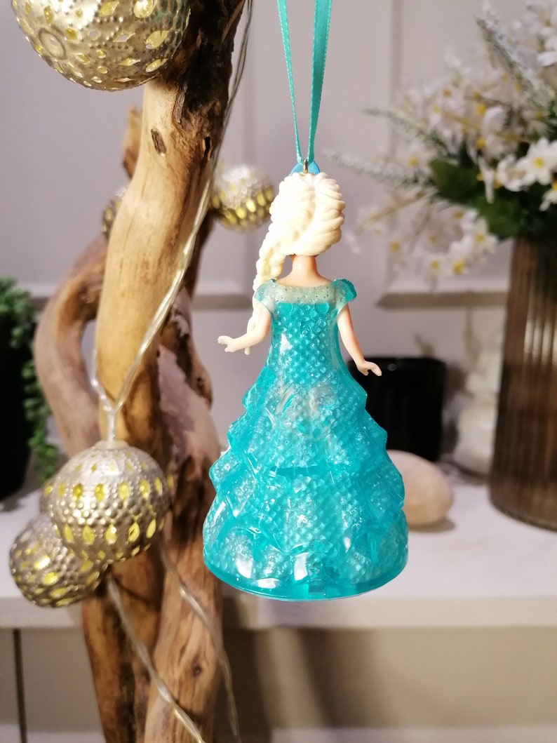 May include: A blue and white plastic ornament of a Disney princess wearing a sparkly blue dress. The ornament is hanging from a blue ribbon.