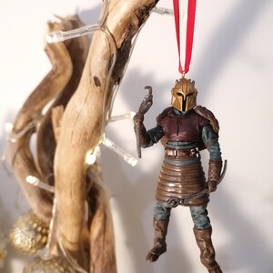 May include: A brown and gold Mandalorian Christmas ornament with a red ribbon. The ornament is hanging from a branch with white lights.