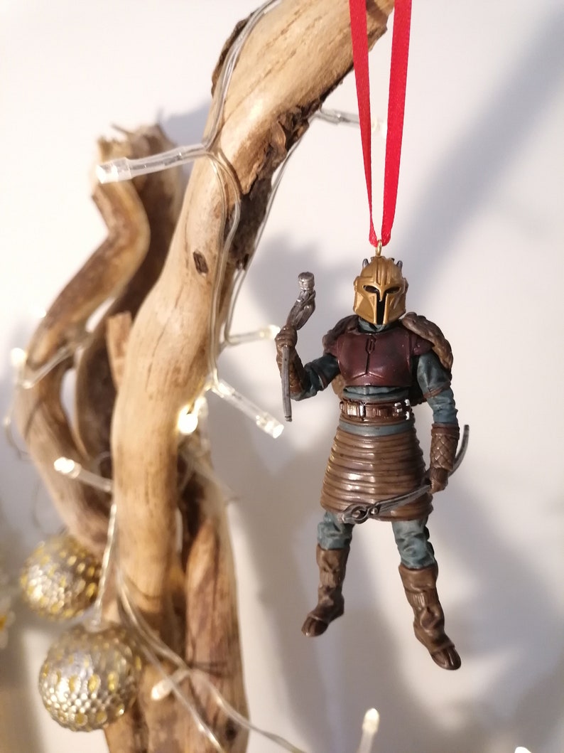 May include: A brown and gold Mandalorian Christmas ornament with a red ribbon. The ornament is hanging from a branch with white lights.