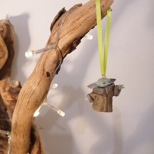 Pu&ograve; includere: Una statuetta verde e marrone di un personaggio del franchise di Star Wars, che indossa un cappotto marrone, appesa a un ramo con un nastro verde.