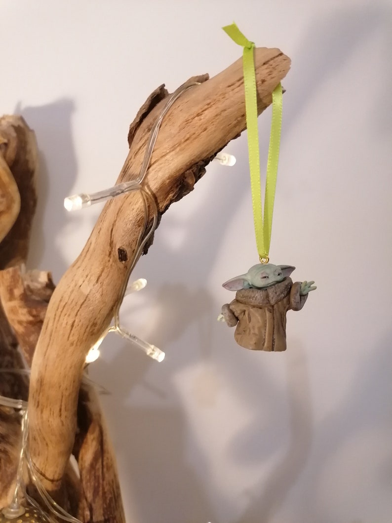 Pu&ograve; includere: Una statuetta verde e marrone di un personaggio del franchise di Star Wars, che indossa un cappotto marrone, appesa a un ramo con un nastro verde.