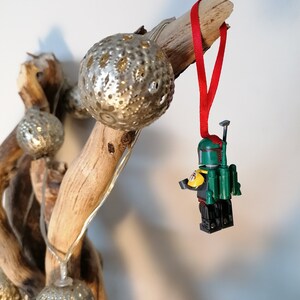 May include: A green and yellow Lego minifigure of Boba Fett hanging from a red ribbon on a driftwood branch with silver ornaments.