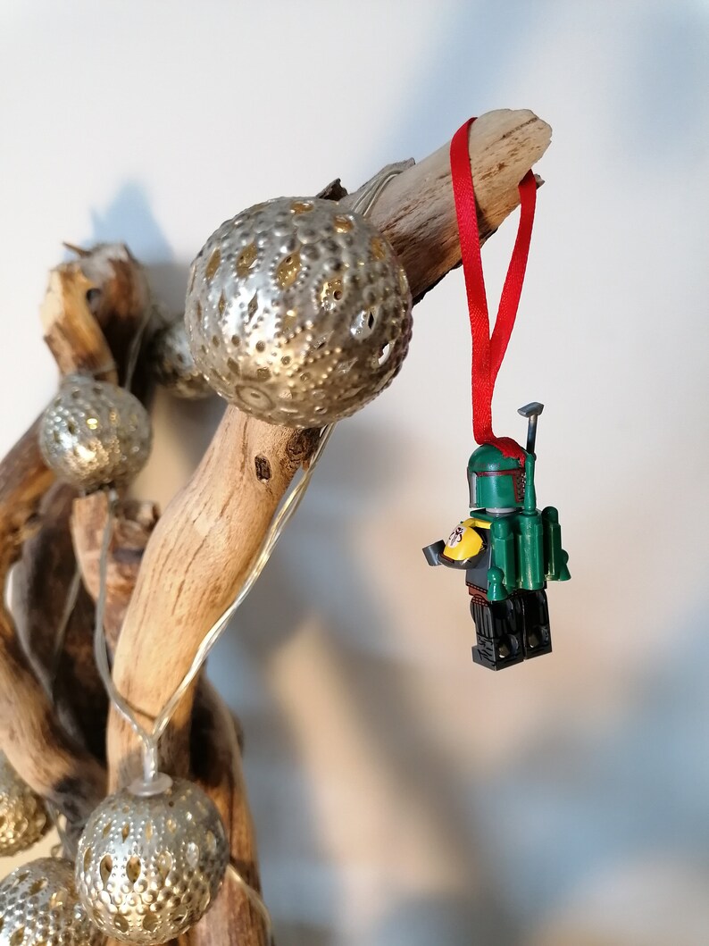 May include: A green and yellow Lego minifigure of Boba Fett hanging from a red ribbon on a driftwood branch with silver ornaments.