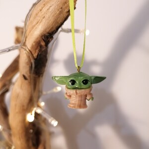 May include: A small, green figurine of a character with large eyes and ears, wearing a tan robe, suspended by a light green ribbon. The figurine is hanging from a piece of driftwood, with small white lights in the background.