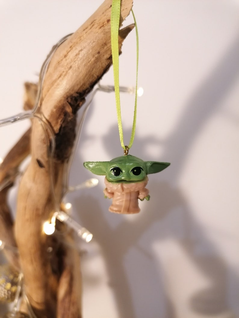 May include: A small, green figurine of a character with large eyes and ears, wearing a tan robe, suspended by a light green ribbon. The figurine is hanging from a piece of driftwood, with small white lights in the background.