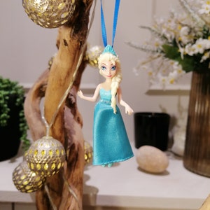 K&ouml;nnte beinhalten: Eine blaue und wei&szlig;e Plastikpuppe mit blonden Haaren, die an einem blauen Band h&auml;ngt. Die Puppe tr&auml;gt ein blaues Kleid und h&auml;lt eine kleine wei&szlig;e Blume.