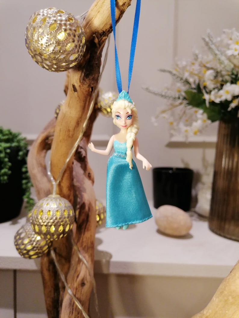 K&ouml;nnte beinhalten: Eine blaue und wei&szlig;e Plastikpuppe mit blonden Haaren, die an einem blauen Band h&auml;ngt. Die Puppe tr&auml;gt ein blaues Kleid und h&auml;lt eine kleine wei&szlig;e Blume.