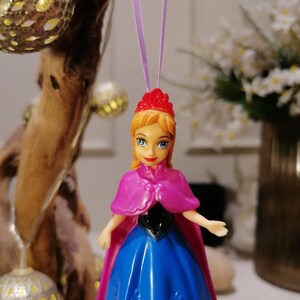 May include: A plastic doll ornament of a princess wearing a blue dress, pink cape, and a red crown. The doll is hanging from a purple string.