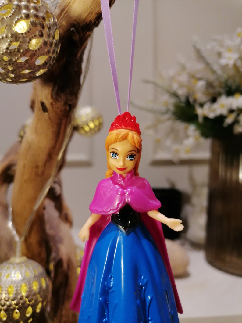 May include: A plastic doll ornament of a princess wearing a blue dress, pink cape, and a red crown. The doll is hanging from a purple string.
