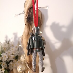 May include: A green and gray Star Wars Boba Fett Christmas ornament with a red ribbon hanging from a branch.