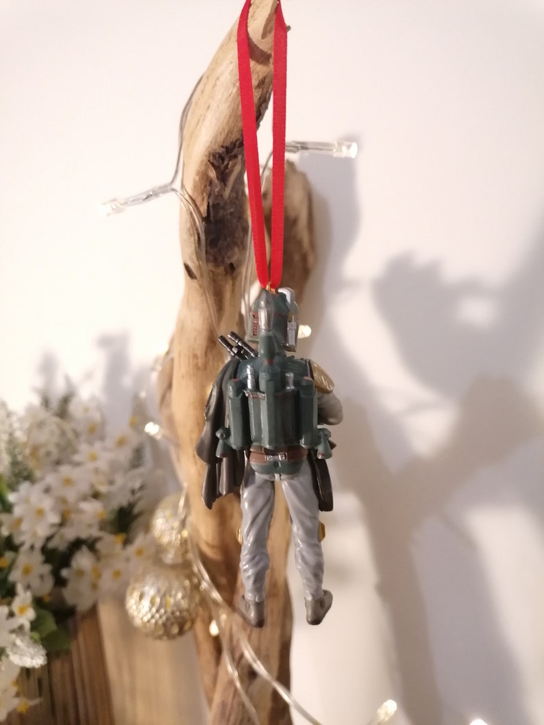 May include: A green and gray Star Wars Boba Fett Christmas ornament with a red ribbon hanging from a branch.