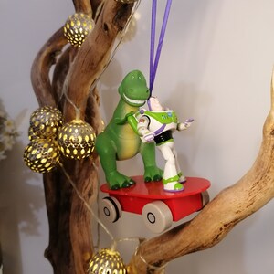 Puede incluir: Adorno de Navidad con los personajes de Toy Story, Buzz Lightyear y Rex, montando un monopatín rojo, colgado de una rama con luces.