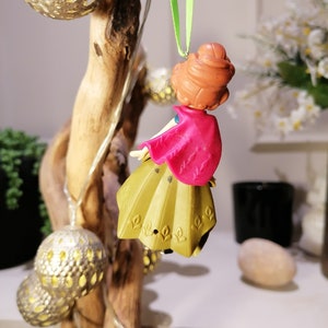 Pu&ograve; includere: Una piccola figurina di plastica di una ragazza con i capelli castani, un mantello rosa e un vestito dorato &egrave; appesa a un ramo con uno spago verde. La figurina indossa uno spago verde intorno al collo.