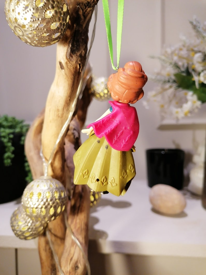 Pu&ograve; includere: Una piccola figurina di plastica di una ragazza con i capelli castani, un mantello rosa e un vestito dorato &egrave; appesa a un ramo con uno spago verde. La figurina indossa uno spago verde intorno al collo.