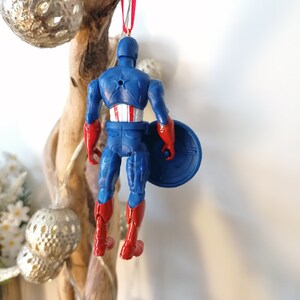 May include: A blue and red plastic Captain America Christmas ornament with a red ribbon and a silver star on the top. The ornament is hanging from a branch.