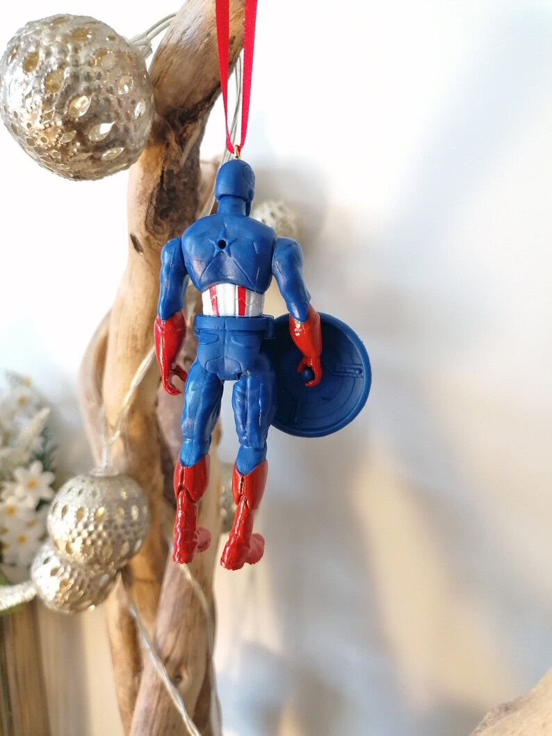 May include: A blue and red plastic Captain America Christmas ornament with a red ribbon and a silver star on the top. The ornament is hanging from a branch.