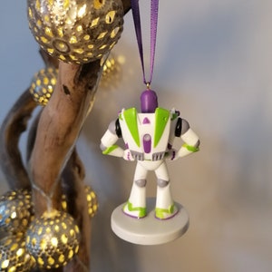 May include: A white and green Buzz Lightyear Christmas ornament with a purple ribbon. The ornament is hanging from a branch with string lights.