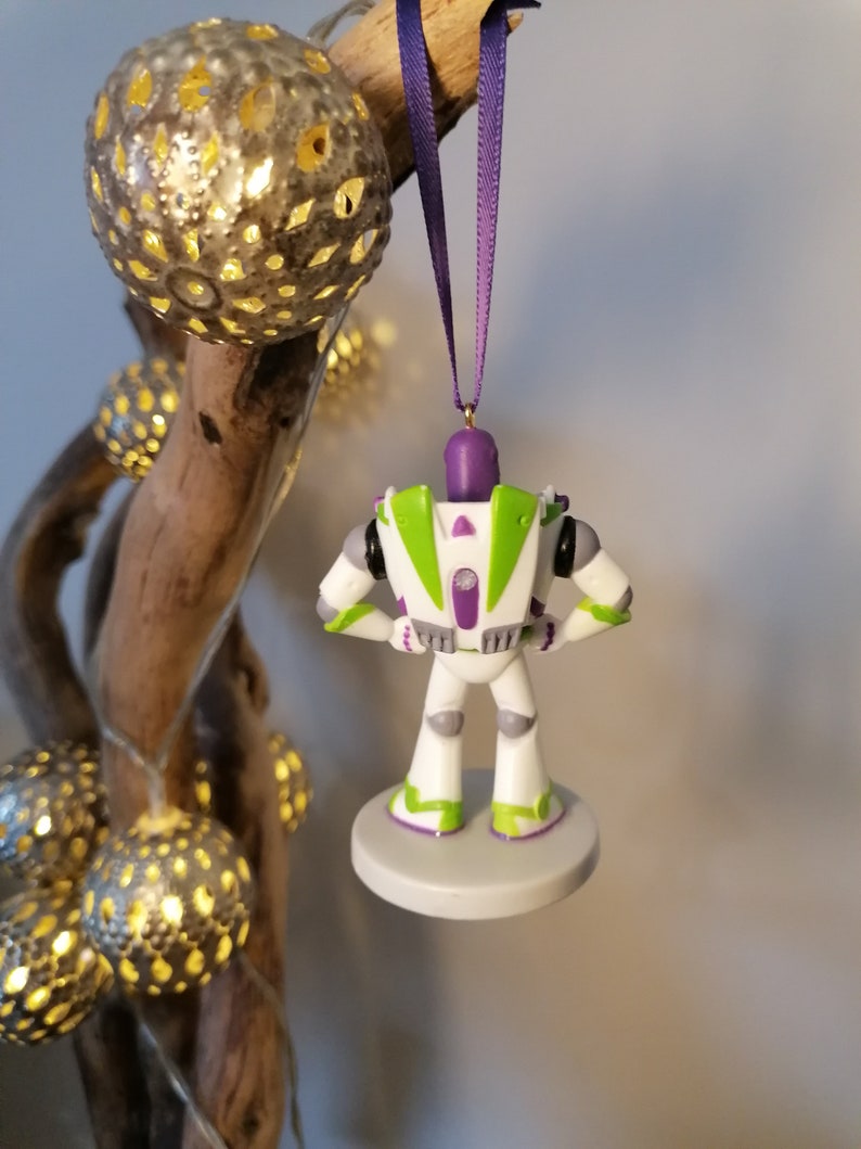 May include: A white and green Buzz Lightyear Christmas ornament with a purple ribbon. The ornament is hanging from a branch with string lights.