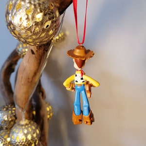 May include: A plastic toy ornament of Woody from Toy Story. Woody is wearing his signature cowboy hat, vest, and jeans. He is holding his trusty sidekick, a brown plastic sheriff's badge.