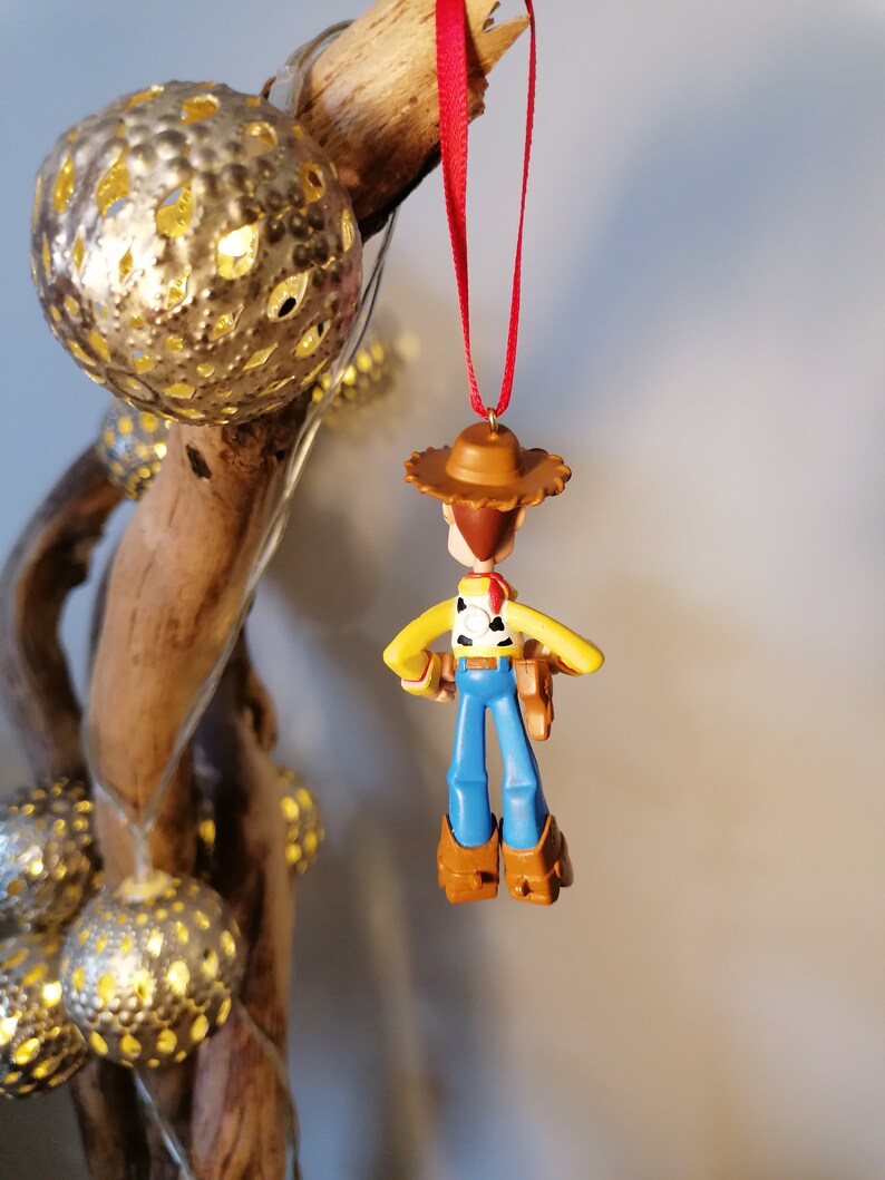 May include: A plastic toy ornament of Woody from Toy Story. Woody is wearing his signature cowboy hat, vest, and jeans. He is holding his trusty sidekick, a brown plastic sheriff's badge.