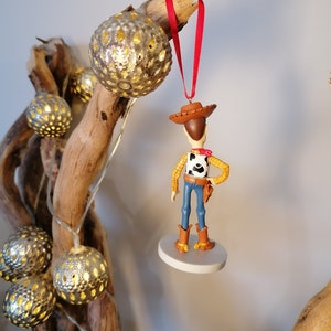 Op de afbeelding: Een rode draad hangt aan een tak met een wit en bruin beeldje van Woody uit Toy Story. Woody draagt zijn kenmerkende cowboyhoed en vest. Het beeldje is bevestigd aan een witte ronde basis.