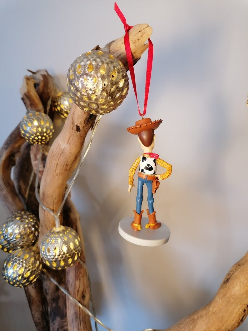 Op de afbeelding: Een rode draad hangt aan een tak met een wit en bruin beeldje van Woody uit Toy Story. Woody draagt zijn kenmerkende cowboyhoed en vest. Het beeldje is bevestigd aan een witte ronde basis.