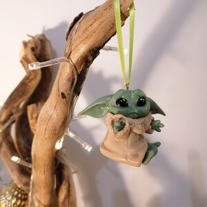 May include: A green and brown figurine of a character from the Star Wars franchise, hanging from a branch with a green ribbon. The figurine is a Christmas ornament.