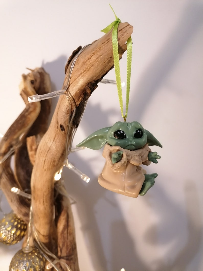 May include: A green and brown figurine of a character from the Star Wars franchise, hanging from a branch with a green ribbon. The figurine is a Christmas ornament.
