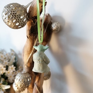 May include: A green Yoda ornament hanging from a branch with a green ribbon. The ornament is made of ceramic and has a detailed design.