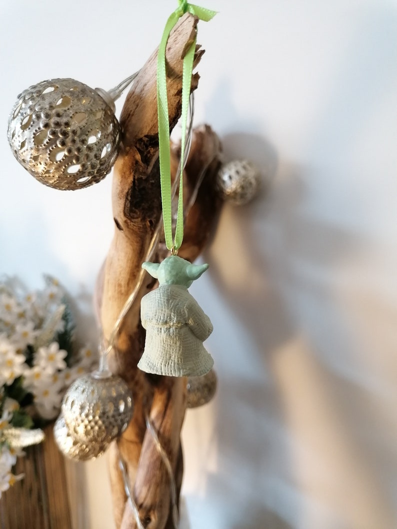 May include: A green Yoda ornament hanging from a branch with a green ribbon. The ornament is made of ceramic and has a detailed design.