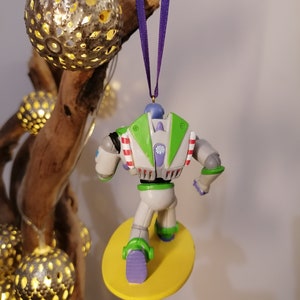 K&ouml;nnte beinhalten: Ein gr&uuml;ner und wei&szlig;er Buzz Lightyear Weihnachtsbaumschmuck mit einem gelben Surfbrett. Der Schmuck h&auml;ngt an einem lila Band.