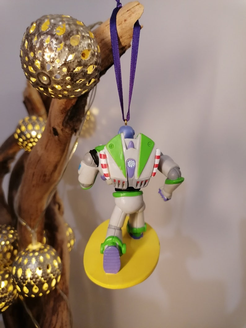 K&ouml;nnte beinhalten: Ein gr&uuml;ner und wei&szlig;er Buzz Lightyear Weihnachtsbaumschmuck mit einem gelben Surfbrett. Der Schmuck h&auml;ngt an einem lila Band.