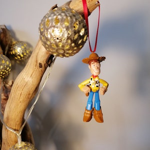 May include: A plastic toy figurine of Woody from Toy Story hanging from a branch with a red ribbon. The figurine is wearing a yellow shirt, blue jeans, a brown hat, and brown boots. The figurine is holding a gun in his right hand. The branch is decorated with gold metallic ornaments.