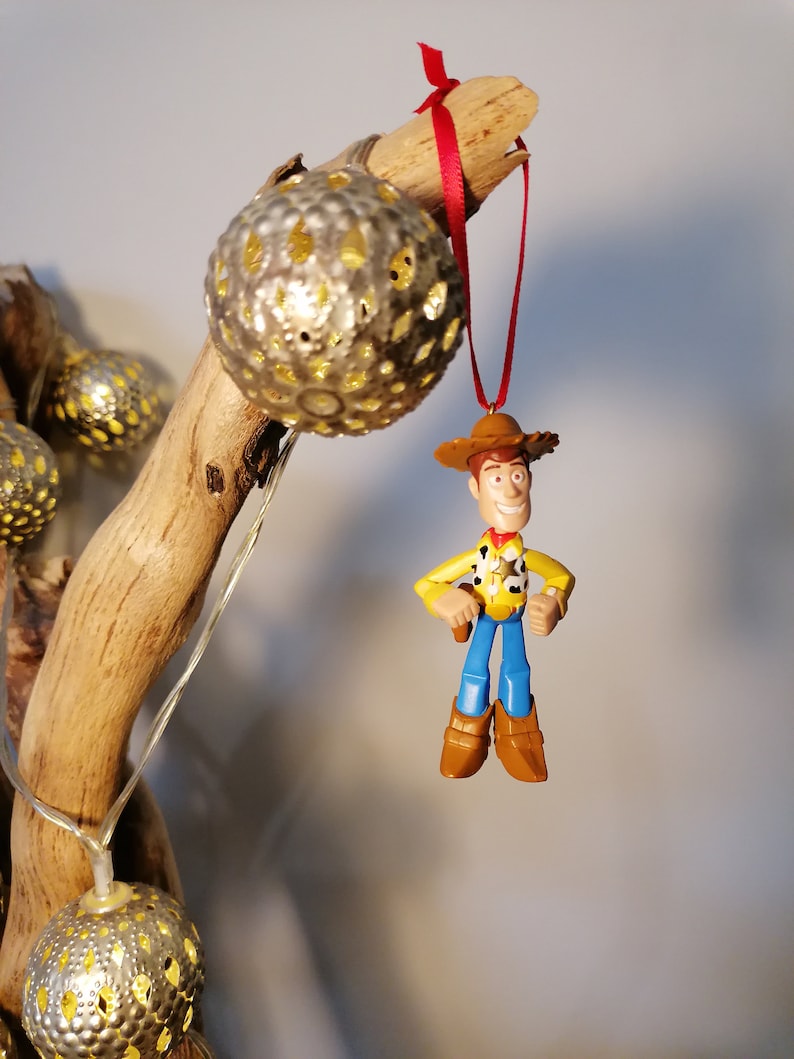 May include: A plastic toy figurine of Woody from Toy Story hanging from a branch with a red ribbon. The figurine is wearing a yellow shirt, blue jeans, a brown hat, and brown boots. The figurine is holding a gun in his right hand. The branch is decorated with gold metallic ornaments.