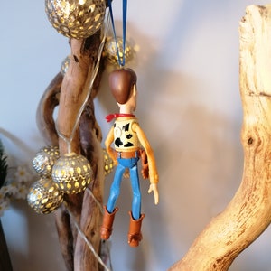Puede incluir: Una figura de juguete de pl&aacute;stico de Woody de Toy Story colgando de una rama con una cuerda azul. La figura lleva su caracter&iacute;stico sombrero de vaquero, chaleco y pantalones vaqueros. La rama est&aacute; decorada con luces de cadena y adornos dorados.