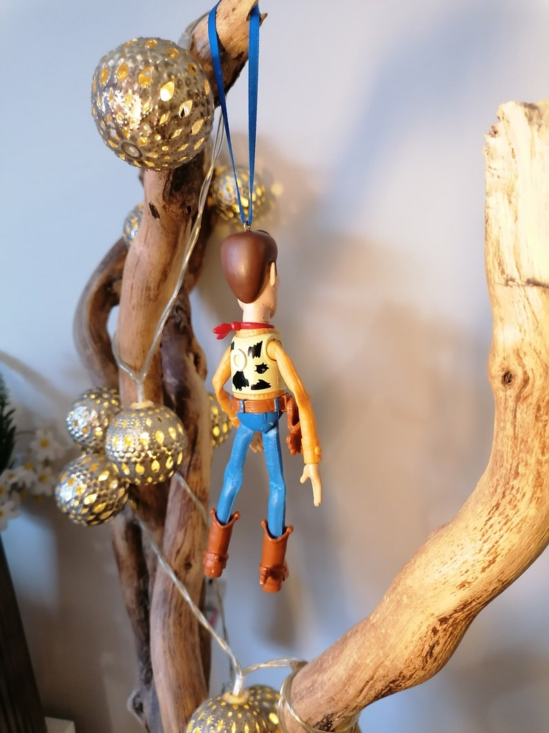 Puede incluir: Una figura de juguete de pl&aacute;stico de Woody de Toy Story colgando de una rama con una cuerda azul. La figura lleva su caracter&iacute;stico sombrero de vaquero, chaleco y pantalones vaqueros. La rama est&aacute; decorada con luces de cadena y adornos dorados.