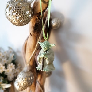 May include: A green Yoda figurine ornament hanging from a driftwood branch with a green ribbon. The ornament is a popular character from the Star Wars franchise.