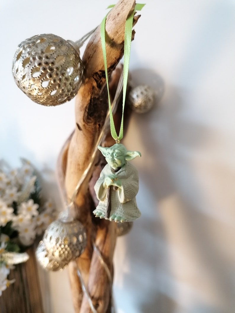 May include: A green Yoda figurine ornament hanging from a driftwood branch with a green ribbon. The ornament is a popular character from the Star Wars franchise.