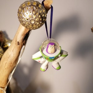 May include: A white and green Buzz Lightyear toy ornament with a purple helmet and a surprised expression. The ornament is hanging from a purple ribbon.