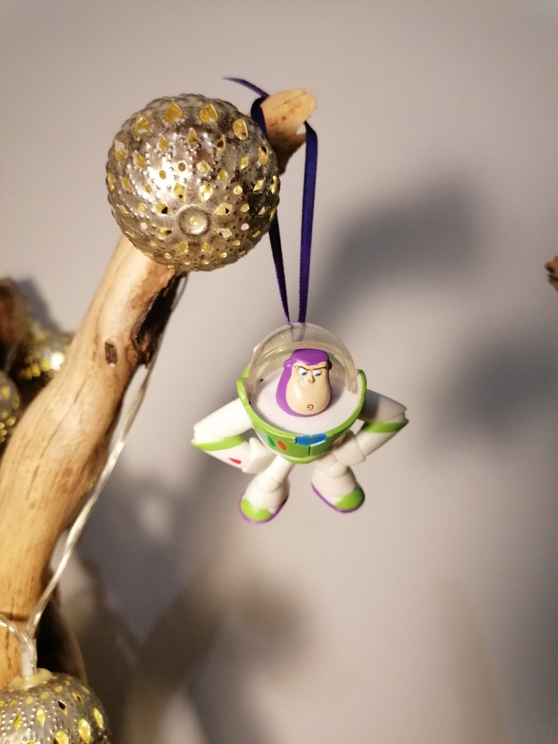 May include: A white and green Buzz Lightyear toy ornament with a purple helmet and a surprised expression. The ornament is hanging from a purple ribbon.