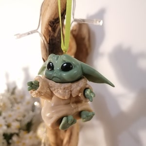 May include: A green and brown plastic ornament of a character from the Star Wars franchise, The Mandalorian. The character is wearing a brown robe and has large eyes. The ornament is hanging from a green ribbon.