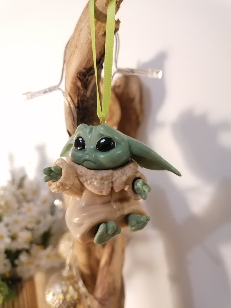 May include: A green and brown plastic ornament of a character from the Star Wars franchise, The Mandalorian. The character is wearing a brown robe and has large eyes. The ornament is hanging from a green ribbon.