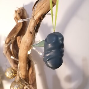 May include: A dark blue, textured, ornament shaped like a bird hanging from a green ribbon on a branch with white lights.