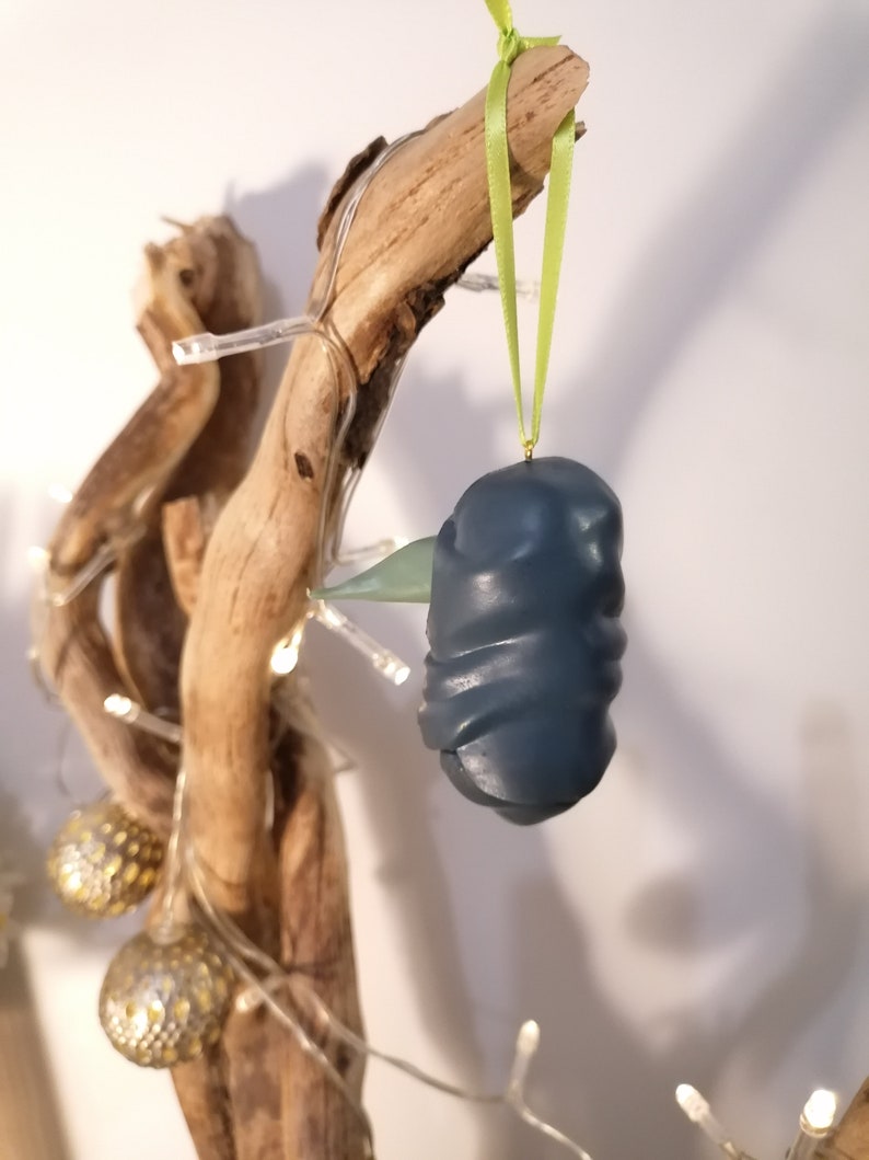 May include: A dark blue, textured, ornament shaped like a bird hanging from a green ribbon on a branch with white lights.