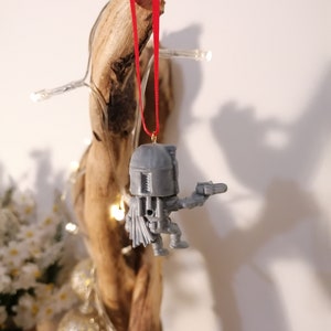May include: A grey plastic ornament of a Star Wars character, Boba Fett, hanging from a red ribbon on a piece of driftwood.
