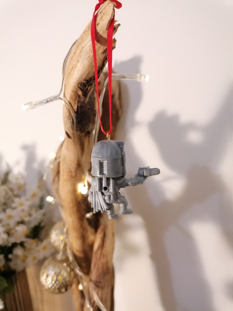 May include: A grey plastic ornament of a Star Wars character, Boba Fett, hanging from a red ribbon on a piece of driftwood.