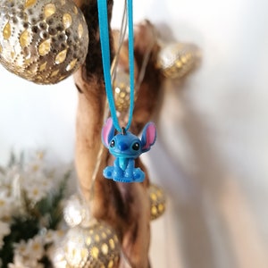 May include: A blue Stitch character charm hanging from a light blue ribbon. The charm is attached to a wooden branch with fairy lights.